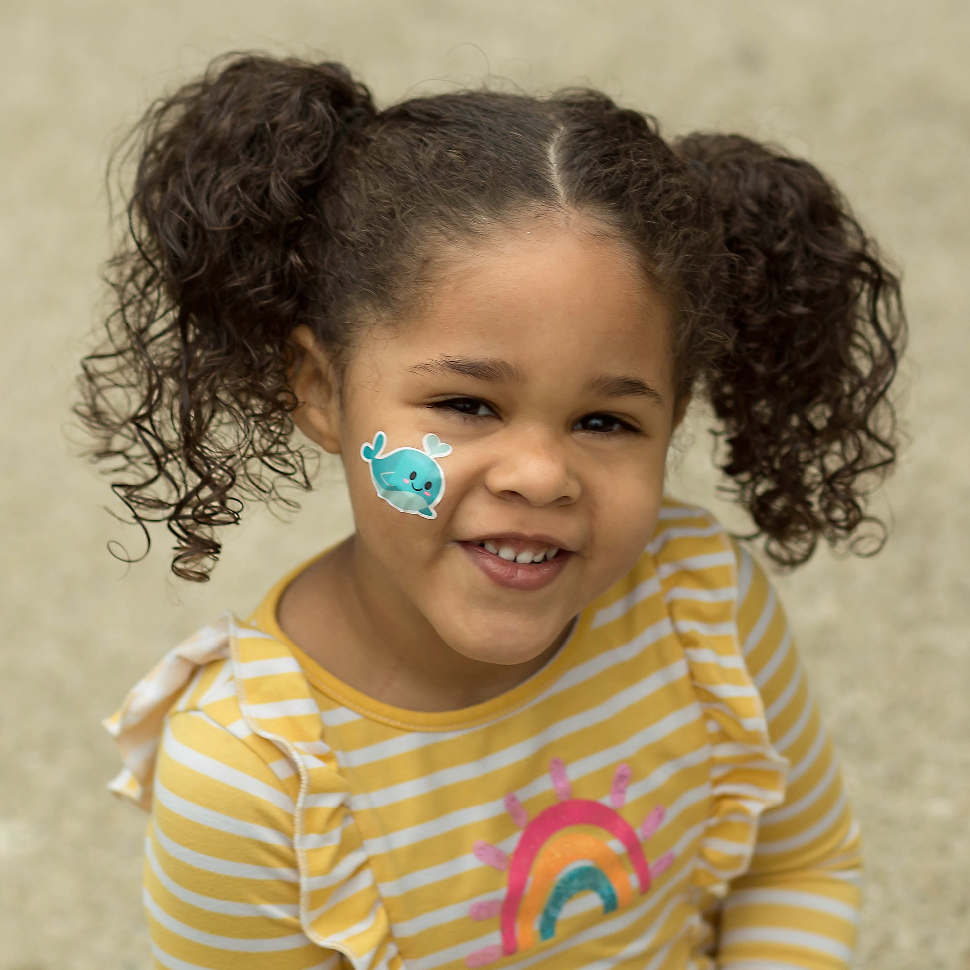 Smiling girl wearing Boo Boo Buddies whale bandage on cheek, adorable waterproof bandaid that brings gentle comfort and cheerful healing.
