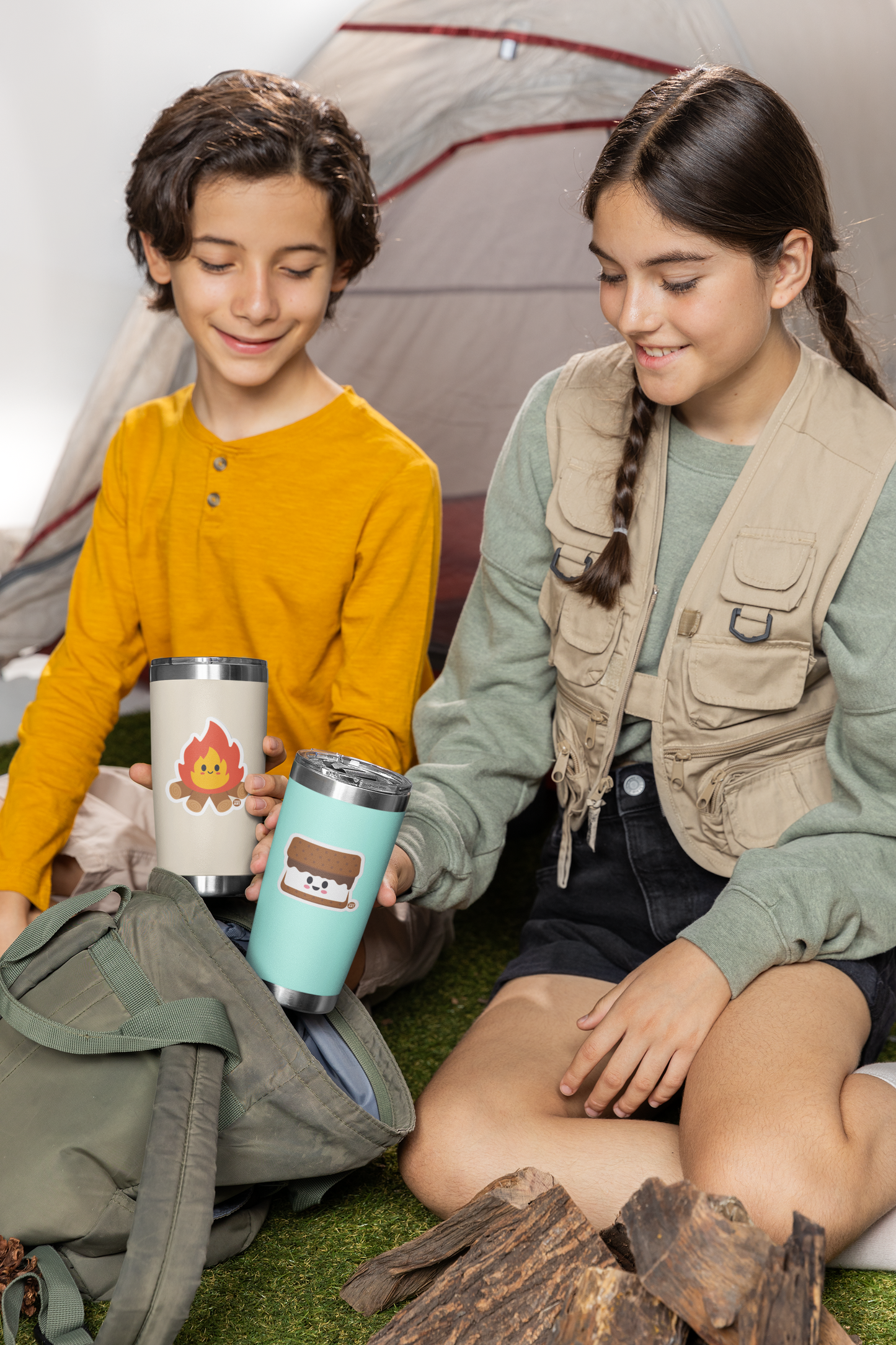 Two children sitting outdoors with camping gear and thermos cups with boo boo buddies stickers.