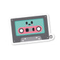 A die-cut vinyl sticker of a cassette tape with a cute character face on it, predominantly teal and pink colors, with a glossy finish.