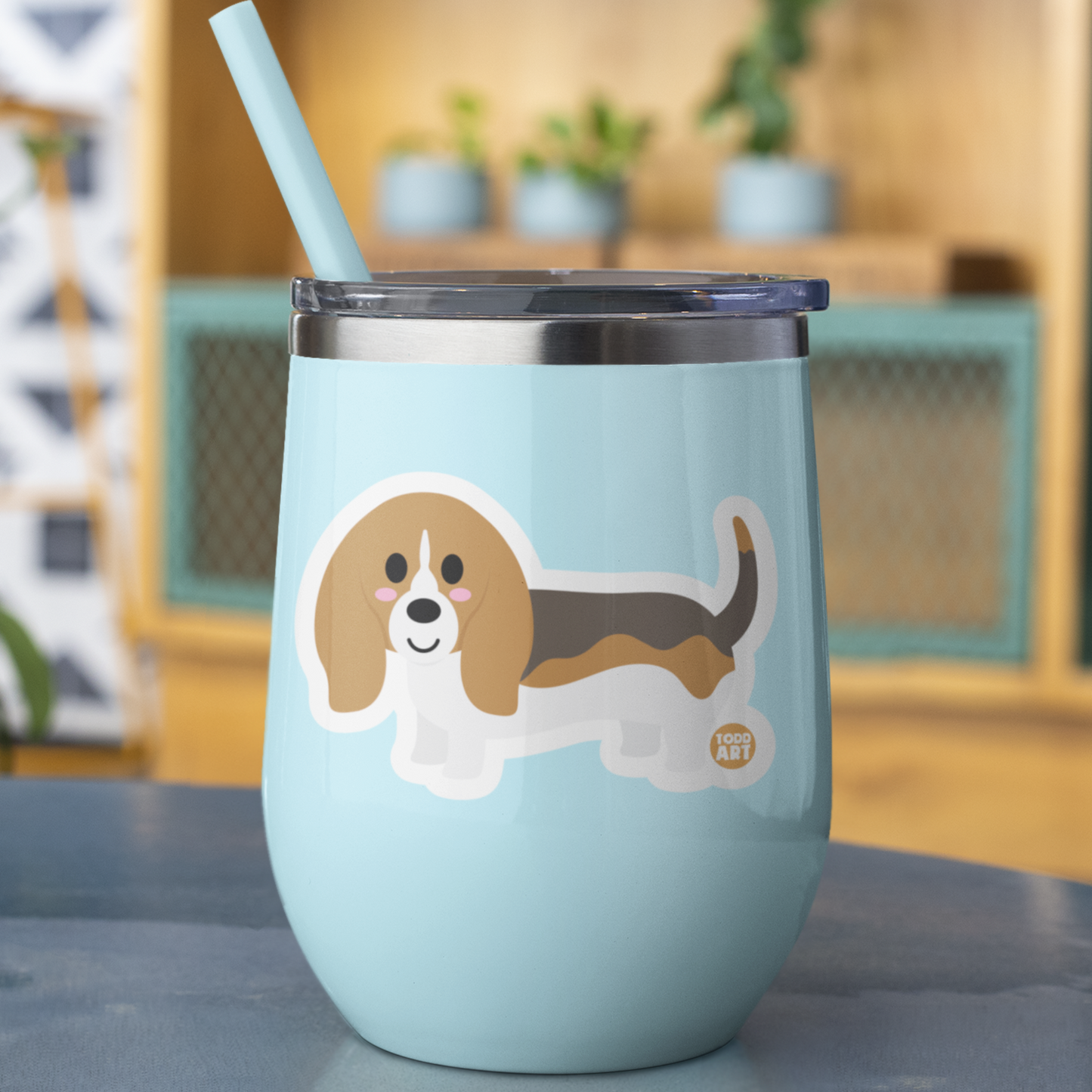 Cute Basset Hound Boo-Boo Buddies® collectible vinyl sticker on a pastel blue tumbler with straw, sitting on a table in a cozy indoor setting
